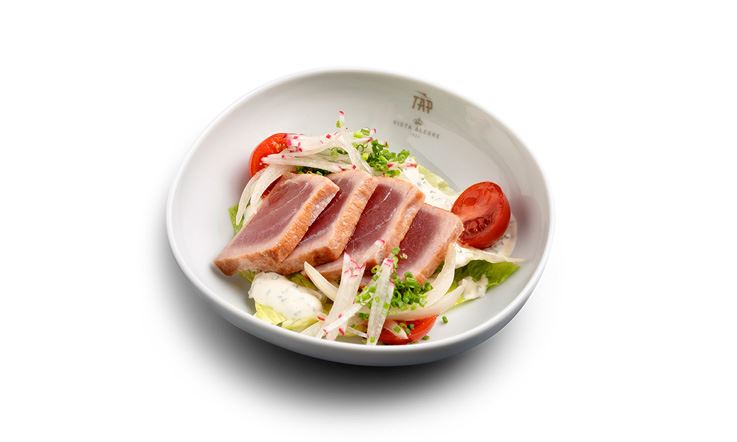 Photography of a deep white plate with the golden TAP Air Portugal logo on the rim, containing rectangular slices of seared tuna, surrounded by green lettuce, red tomatoes cut in half and white strips of Setúbal onion.